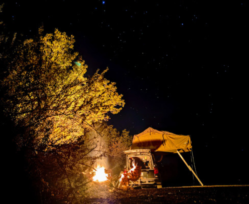 A person sits by a campfire next to a vehicle with a rooftop tent, surrounded by trees under a starry night sky.